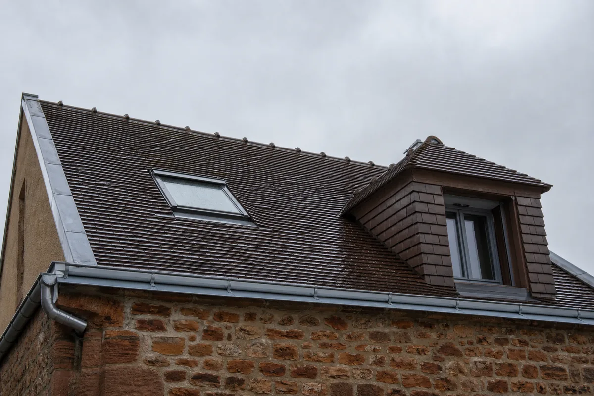 Toiture de faubourg manceau en tuile brune avec une fenêtre de toit et une lucarne, sous un ciel gris perle
